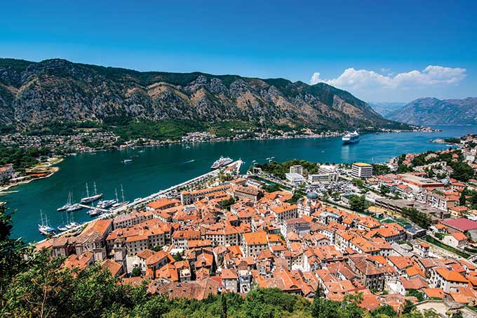 Aerial view of a coastal town with red-roofed buildings, surrounded by mountains and blue water.